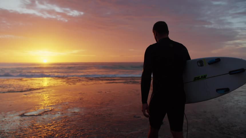A man with a surfboard walks along the wet shoreline at sunset by the ocean