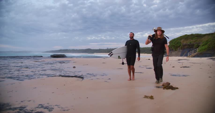 Two men walk across the beach sand at middayone in a swimsuit carrying a surfboard the other holding a tool