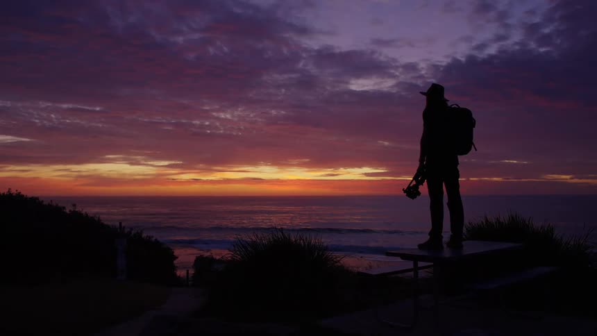 A silhouetted photographer stands atop a picnic table overlooking the coast holding a tripod against a backdrop of a vibrant purple and orange sunset