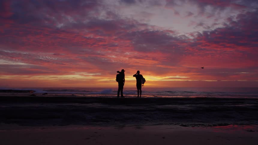 A man holding a surfboard and a man carrying a camera stand silhouetted on a beach observing the waves at a dramatic red sunset