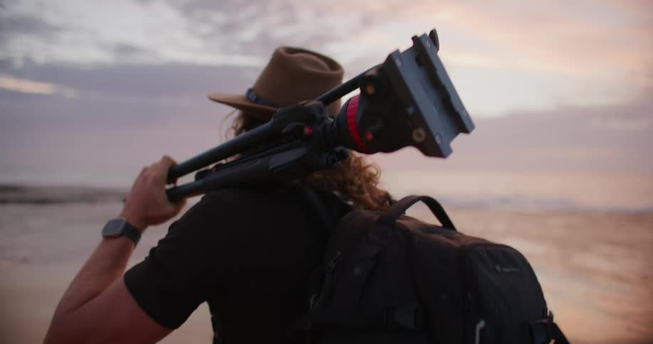 Man carries his camera on a tripod over his shoulder as he walks along the coast