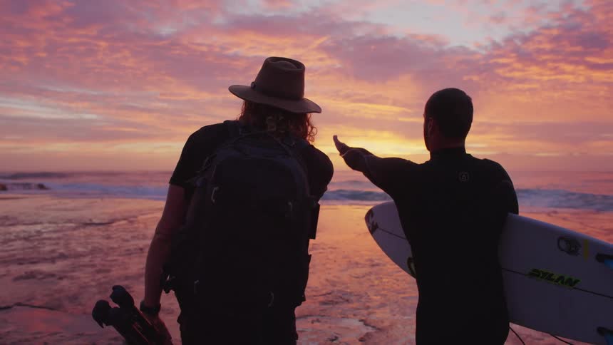 Silhouetted against a vibrant pastel sunset a photographer and a surfer stand on the rocky shoreline gazing out at the ocean and pointing toward the horizon as they scout the waves