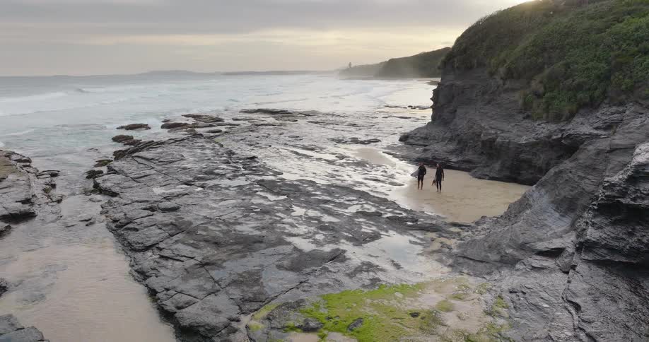 Two people walk along a rocky coastal shoreline beneath an overcast sky