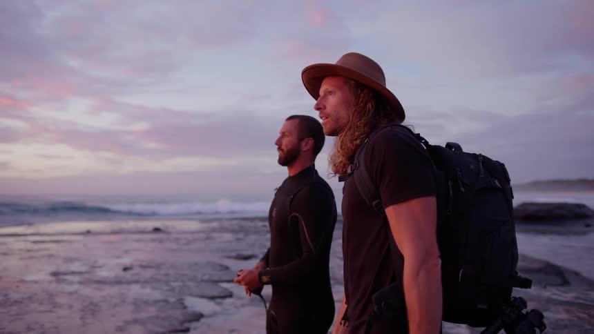 Two men stand along the coast talking and pointing as waves roll in to shore