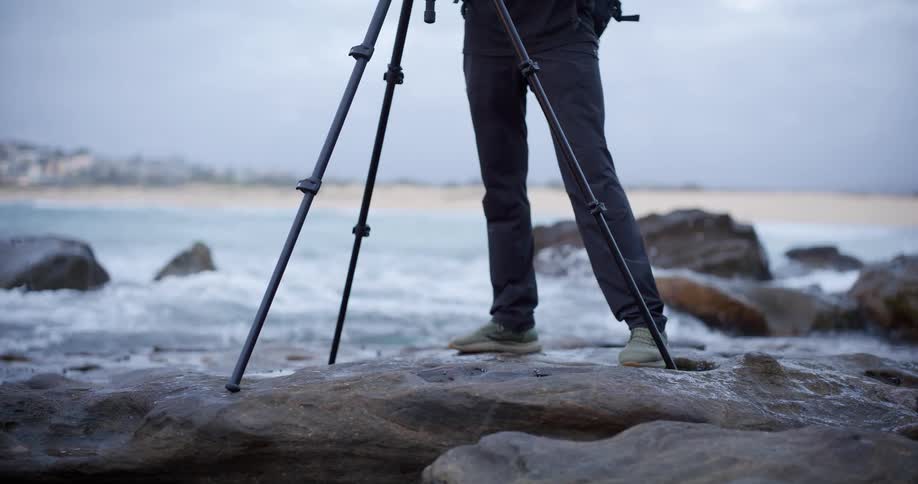 A low shot shows the legs of a person standing firmly on a wet dark rock near the ocean adjusting a black tripod while small waves gently break on the nearby sandy shoreline and background rocks