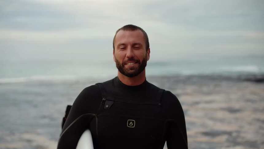 A man in a wet suit smiles as he holds his surfboard while at the ocean