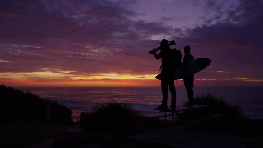 Two men stand on a cliff overlooking the ocean at sunset one with a surfboard and the other with a camera