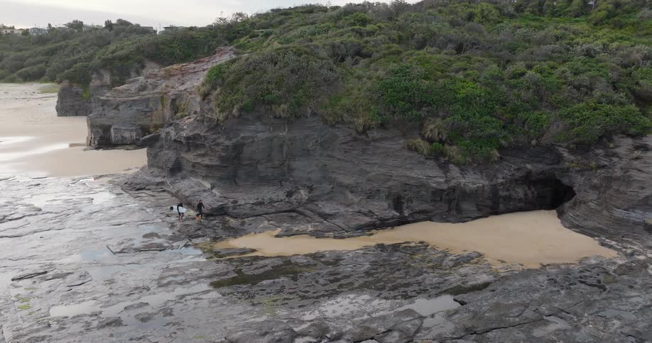 Two people walk near a cliff along the coast as a man carries his surfboard