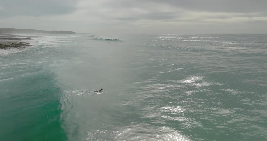 Surfer lies on his surfboard and paddles as waves roll in to shore