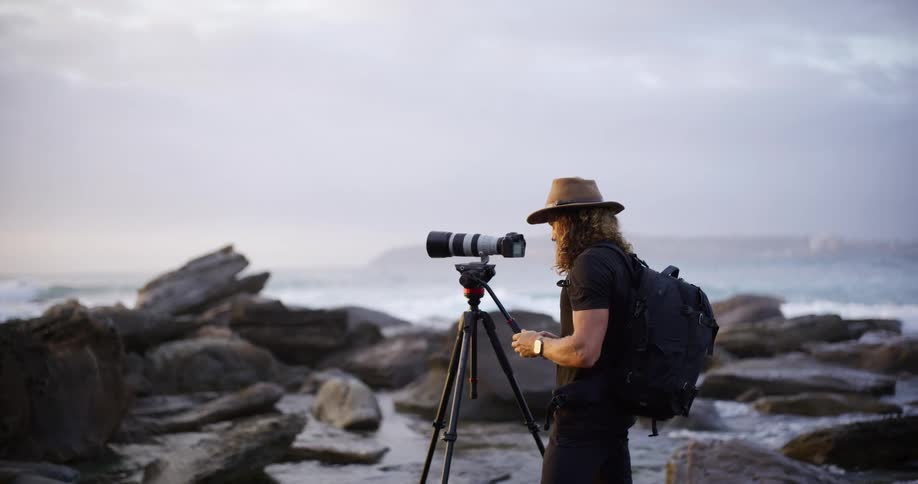A photographer wearing a widebrimmed hat and backpack is standing on a rocky shoreline operating a camera mounted on a tripod with a long lens the setting is overcast with crashing waves visible in the background suggesting the photographer is capturi