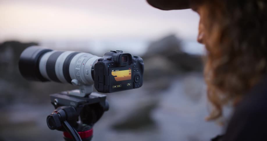 A shallow depthoffield closeup shot focuses on the back of a mirrorless camera mounted on a tripod with a large telephoto lens capturing a person looking over their shoulder at the live view screen in soft cloudy twilight conditions