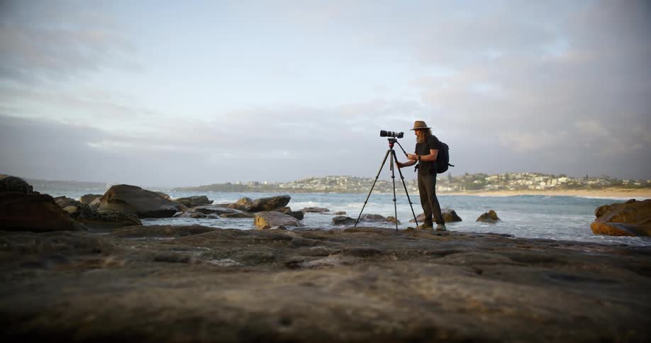 A person wearing a hat and carrying a backpack is standing on a rocky shoreline operating a camera mounted on a tall tripod in the background across the water a town or coastal settlement with buildings is visible under a slightly overcast sky