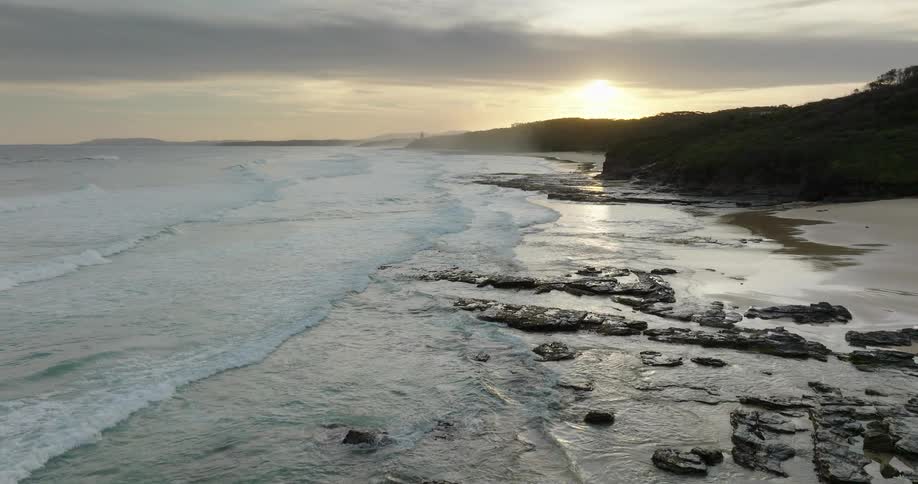 An aerial shot glides over jagged coastal rocks and crashing waves revealing a pristine sandy beach under the warm glow of a setting sun