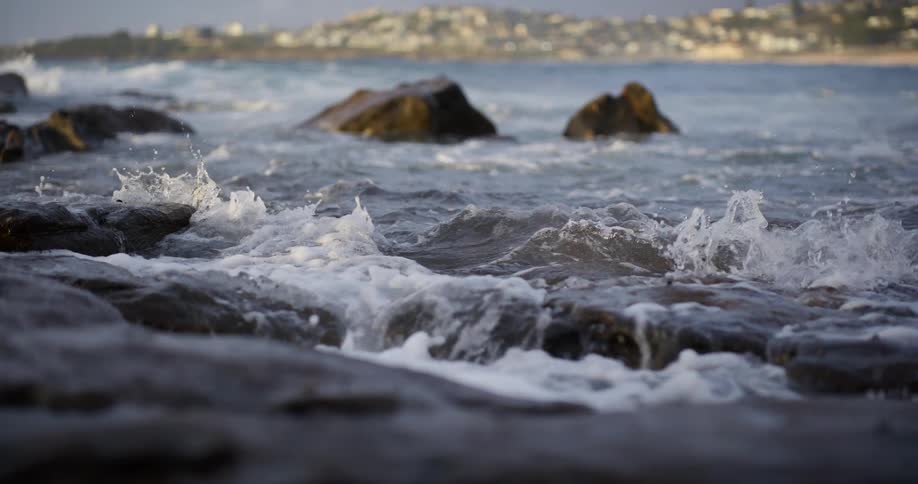 Ocean waves crash dramatically onto dark coastal rocks with a bright distant cityscape visible in the background conveying a feeling of dynamic untamed nature