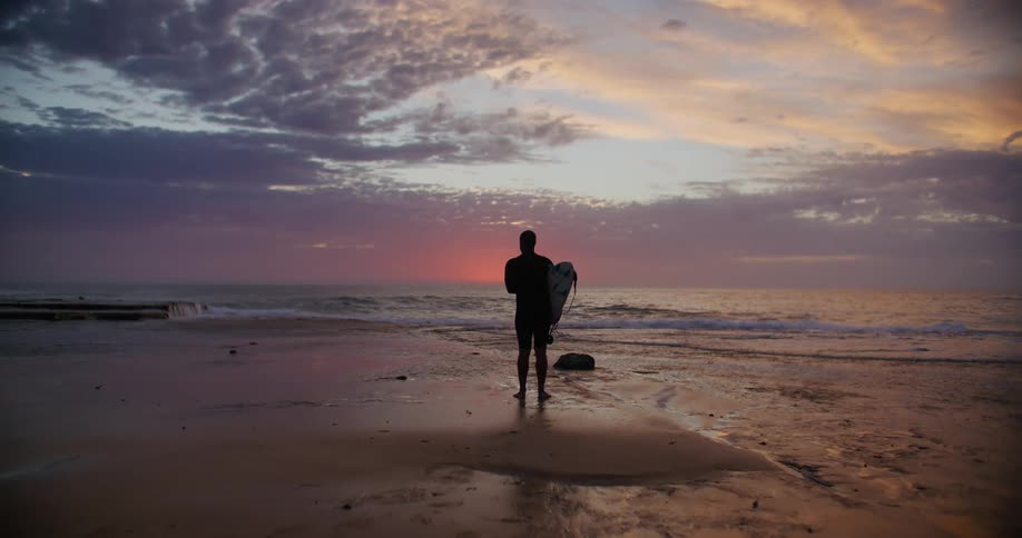 Man stands on the beach holding his surfboard as waves roll in to shore under a cloudy sky