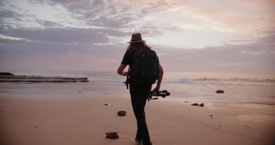 A person runs along the sandy beach at sunset carrying camera equipment