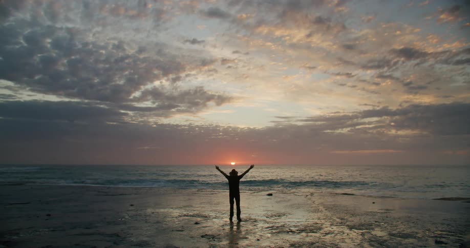 Person with raised arms on wet sand at sunset with illuminated clouds on the horizon