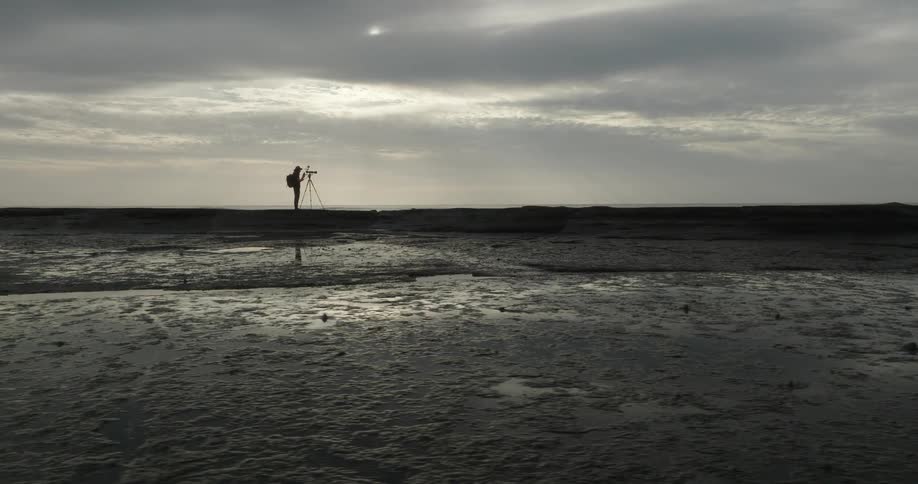 The photographer is silhouetted against the bright cloudy sky and is positioned behind a camera on a tripod seemingly taking a picture of the scene the foreground is a vast reflective stretch of dark wet textured ground possibly mudflats or a beach