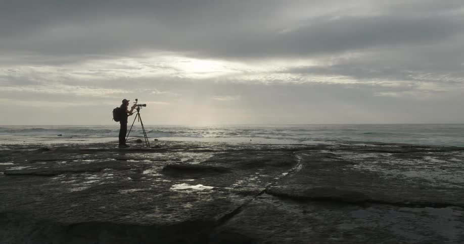 A person stands on a rocky shore recording the ocean as waves roll in beneath cloudy skies