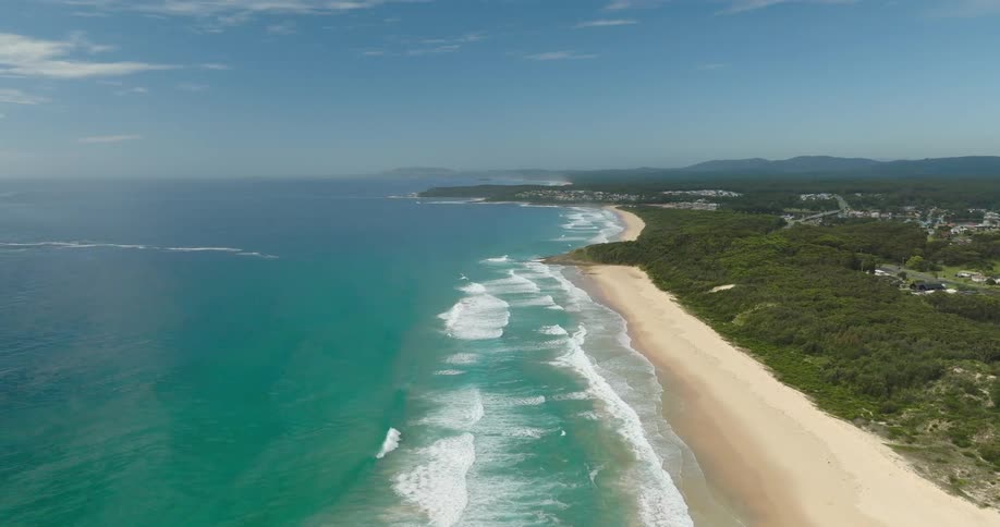 An expansive aerial view captures a beautiful coastline where turquoise ocean waves crash onto a long stretch of golden sand the beach is backed by a dense green forest leading to a small residential area in the distance under a bright blue sky