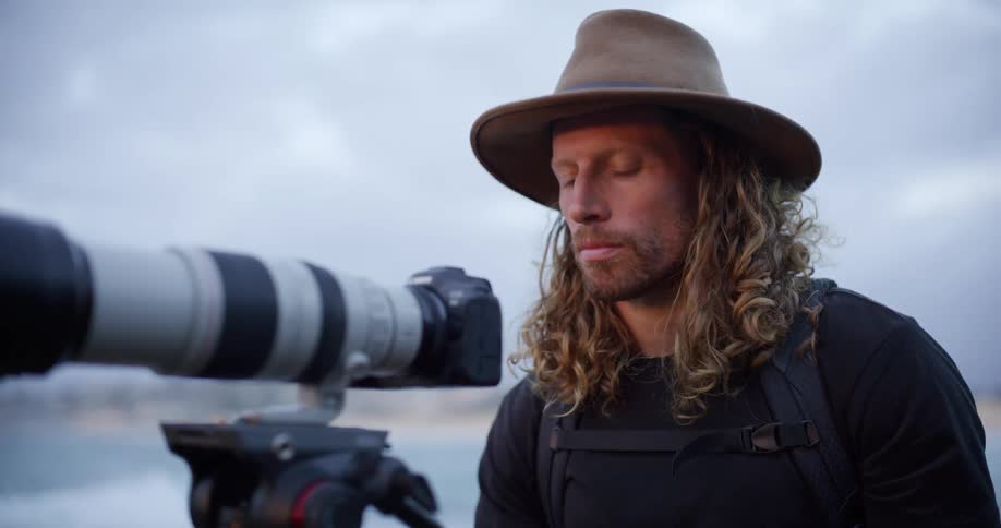 A white man appearing focused and intense is looking forward over the ocean while wearing a fedora and standing next to a professional camera with a long lens on a tripod