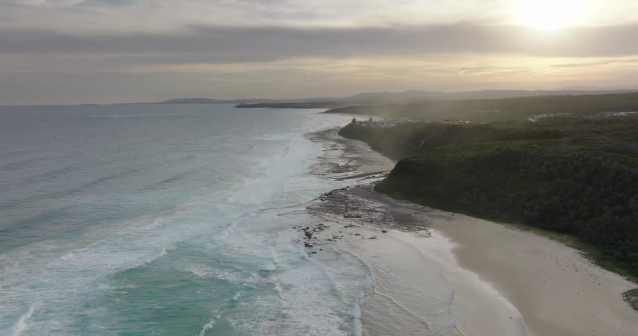 An aerial view of a stunning coastal landscape shows white waves crashing onto a wide sandy beach backed by dark forested cliffs under a golden hazy sky