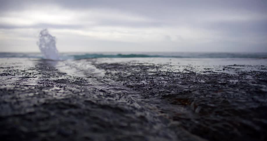 Wave hitting wet rocks on ocean shore under cloudy sky at dusk