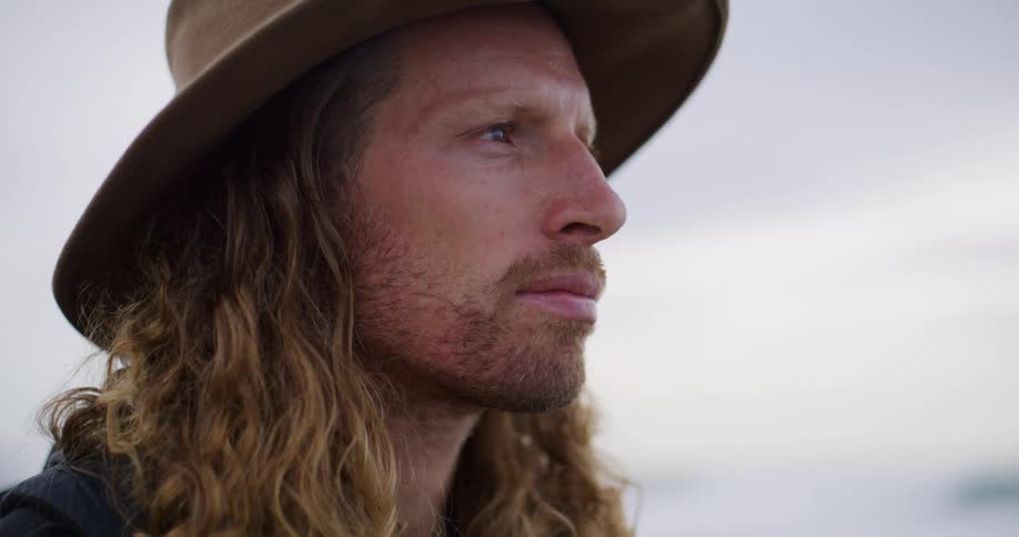 A man with long hair and a hat gazes thoughtfully into the distance