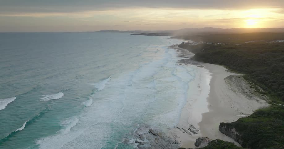 An aerial view reveals waves crashing on a sandy beach alongside lush cliffs under a golden sunset