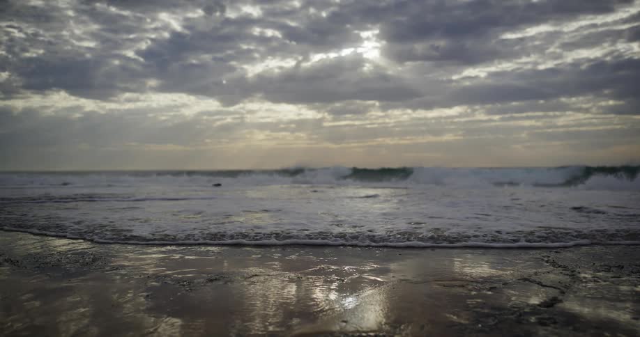 A gentle ocean wave rolls ashore in slow motion its foam washing over the wet sand as moody clouds drift across the sky above