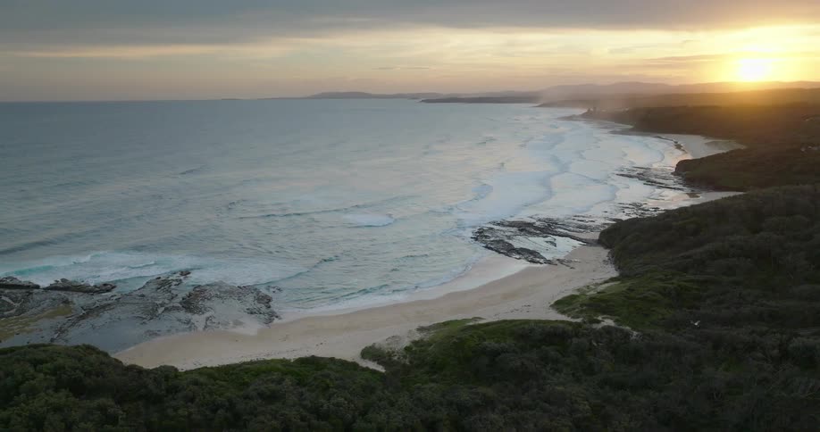 Aerial view of waves crashing on a rocky shore with coastal greenery at sunset