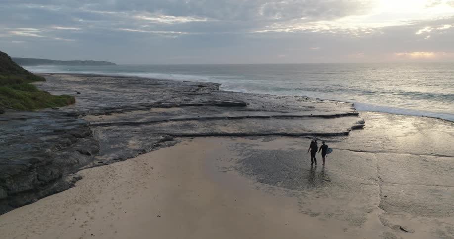 Two people walking on the sand near rocky shores at sunrise