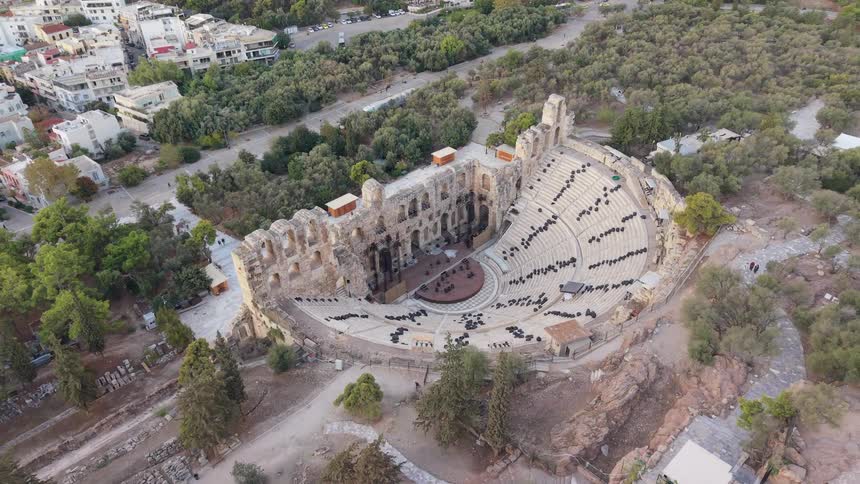 An aerial shot slowly orbits a magnificent ancient stone amphitheater where an orchestra is seated on the stage and throughout the historic stone tiers