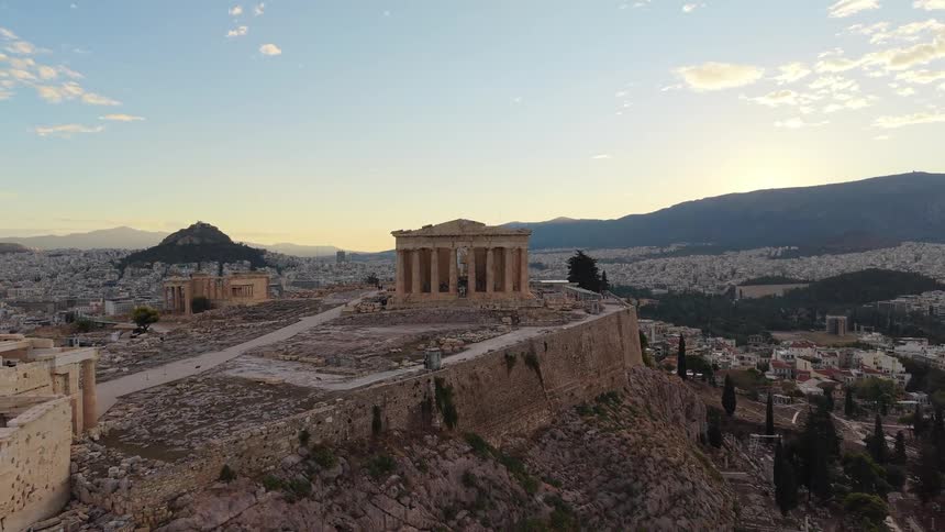 An aerial view shows the ancient greek temples of the acropolis on a rocky hill overlooking the vast city of athens at sunset under a bright sky with scattered clouds