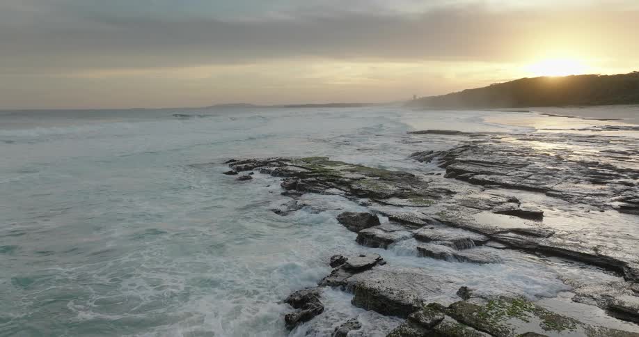 Aerial view of a rocky shoreline with waves crashing under a cloudy sunset sky