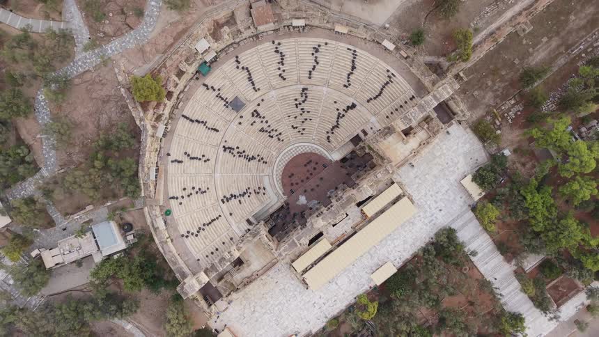A slow rising aerial shot captures a large crowd of people scattered across the weathered stone tiers of an ancient amphitheater