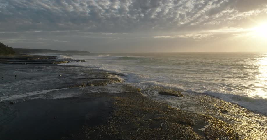 The ocean waves roll continuously onto a wet dark rock shelf reflecting the bright golden sunlight under a sky filled with patterned clouds