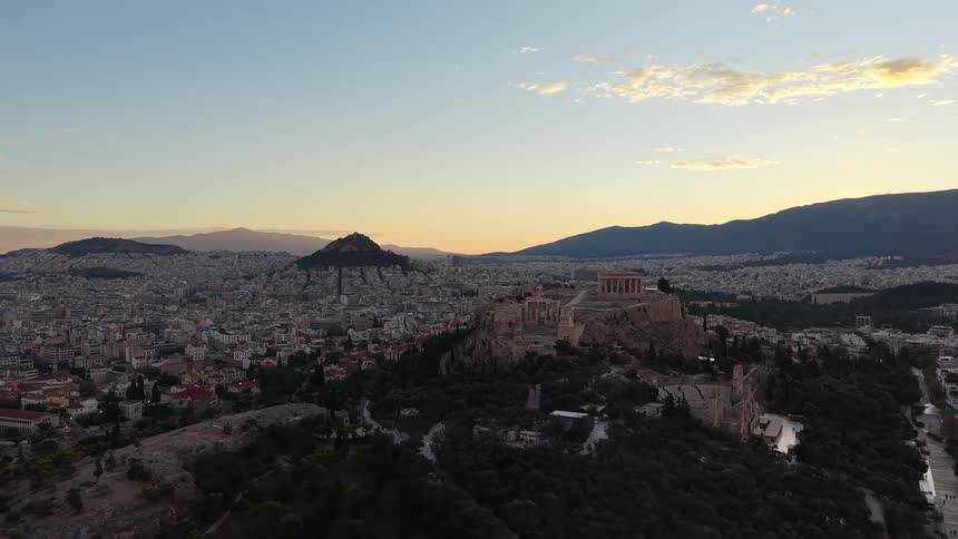 An aerial shot pulls back from ancient historical buildings atop a hill at sunset revealing the vast city skyline and distant mountains bathed in golden light