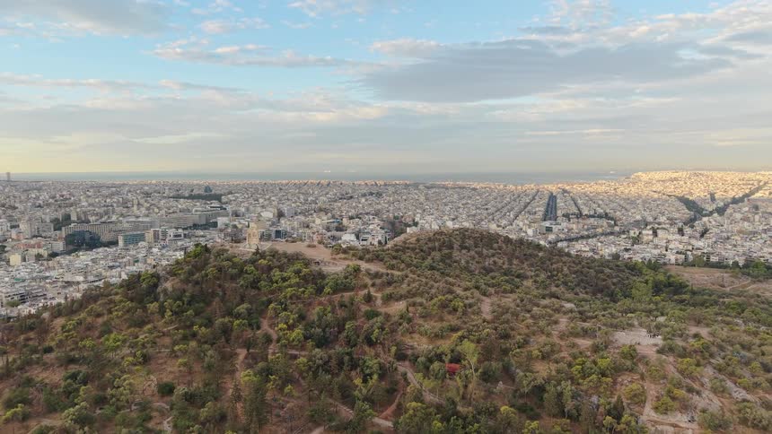 An elevated wide shot captures a large treecovered arid hill sloping down toward the city of athens which spreads out into a massive dense sprawl of white buildings reaching the distant horizon under a blue sky