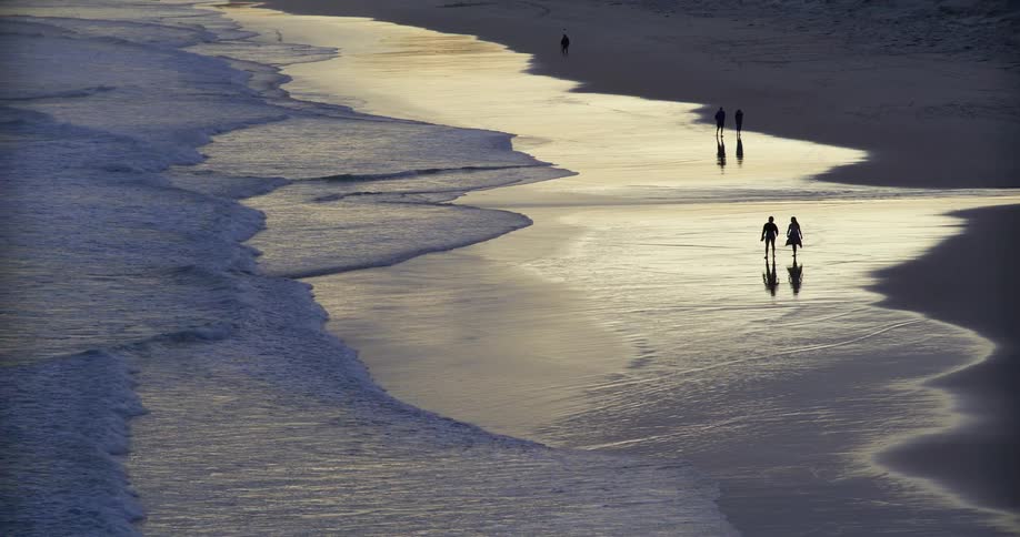 People walking on the sand strip reflected by the low light beside gentle waves