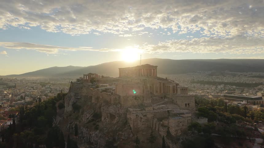 An aerial view of the acropolis of athens featuring the parthenon and surrounding structures perched atop a rocky hill with the city of athens sprawling in the background under a bright sun