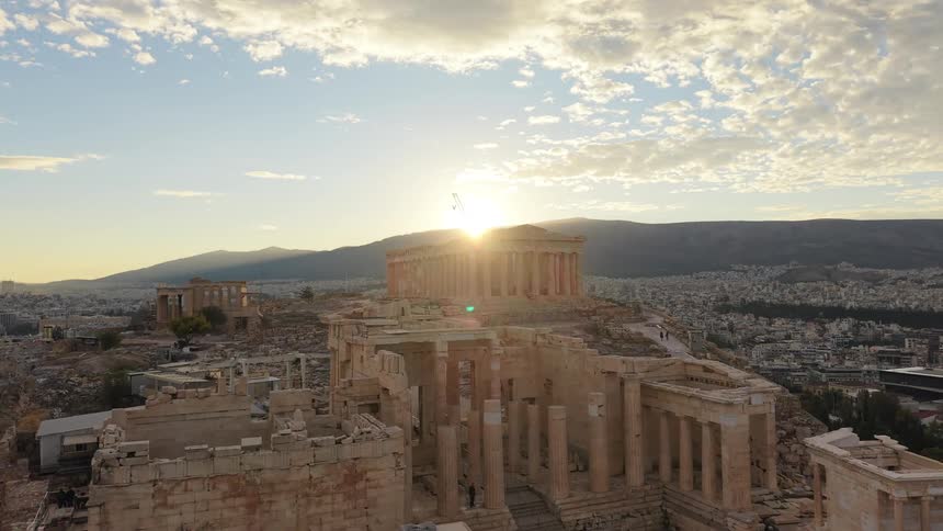 An aerial shot captures the ancient acropolis ruins and the parthenon temple at golden hour overlooking the extensive city of athens with a bright sun flare above the distant mountains