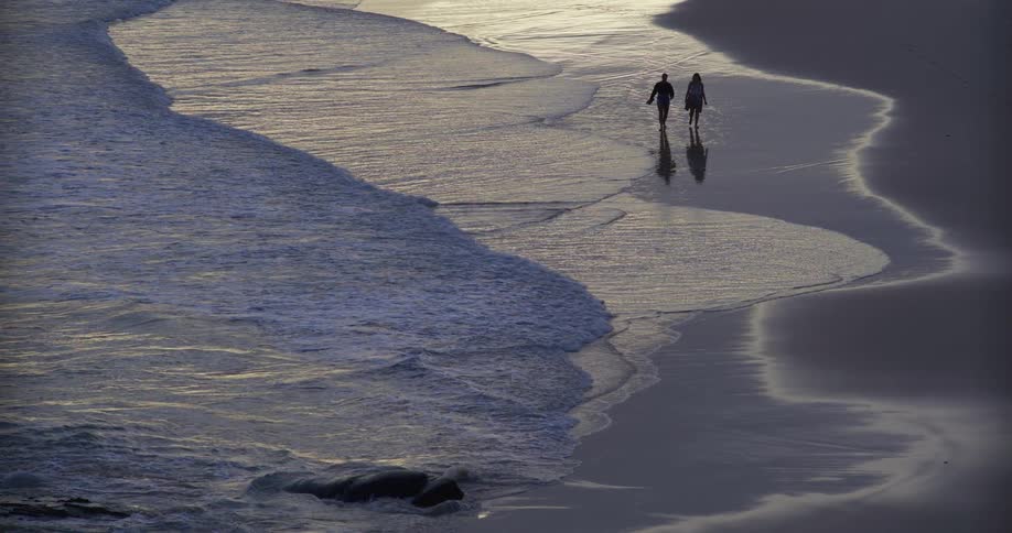 Two people walk along the beach as waves gently hit the shoreline at sunset