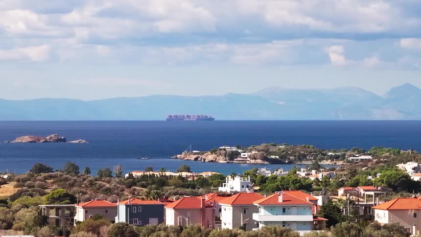 Houses and buildings in a neighborhood along the coast as a boat filled with shipping containers moves across the water
