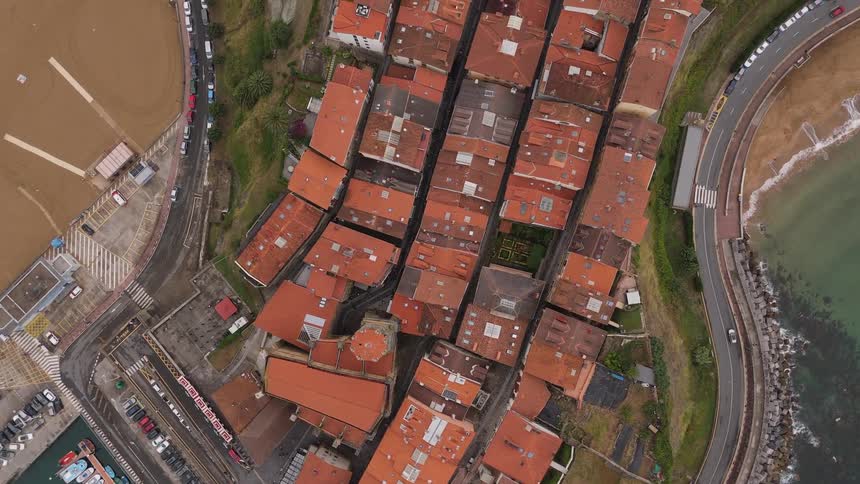The video is an aerial vertical shot that zooms out from a dense coastal townthe view features terracottaroofed buildings clustered together bordered by a busy marina roads grassy hillsides and a sandy beach meeting the ocean