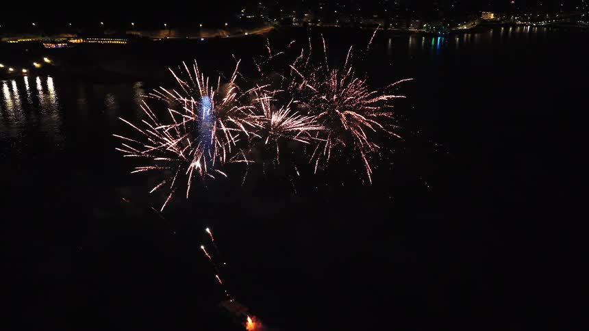 A nighttime fireworks display over a body of water likely a lake or bay with the bright sparks reflecting on the dark surfacethe fireworks primarily white and golden bursts are launched from a small platform on the water with the distant shore lig