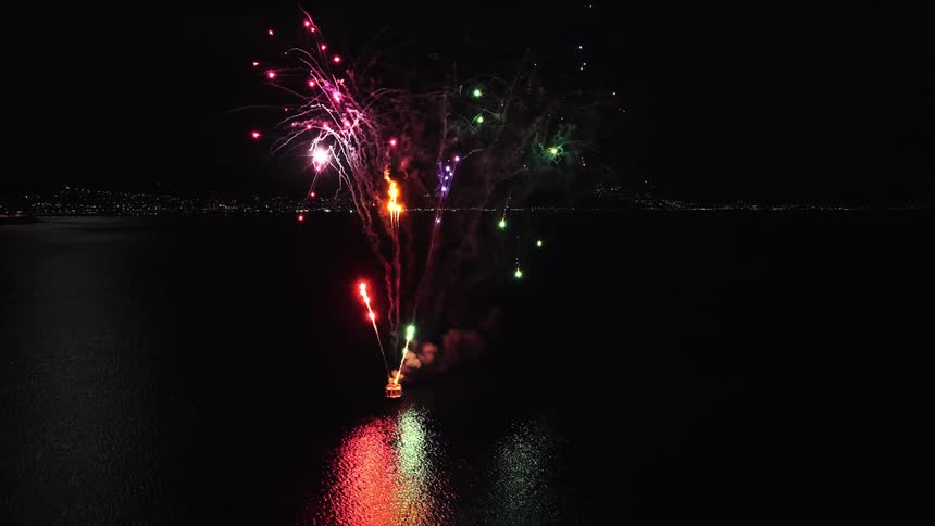 Fireworks launched from a boat on the water glow as they explode and leave smoke trails in the night sky along the coast