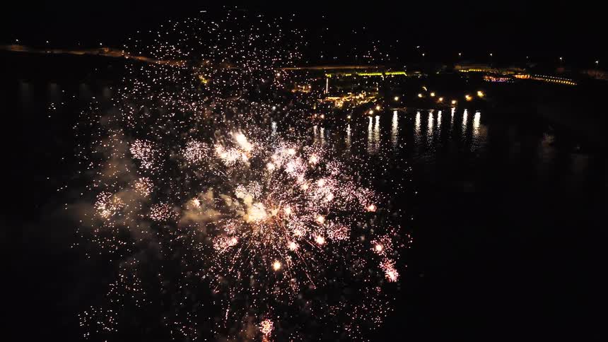 Fireworks show over water at night with bright reflections and lit cityscape in the background