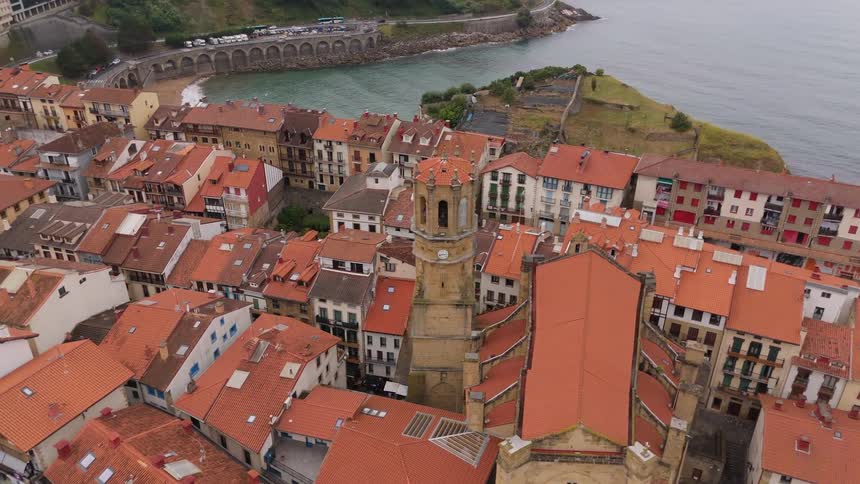 This is a highangle vertical flyover shot of a coastal town centered on residential buildings with terracotta roofsthe camera tracks slowly over the dense urban structure showing the proximity of the town center to a marina a busy road running alo