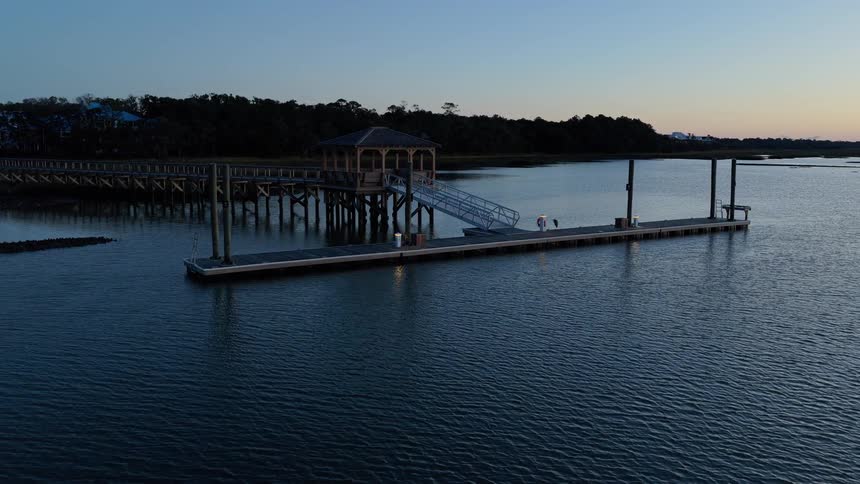 Wooden pier with covered structure extends over calm water at dusk surrounded by vegetation